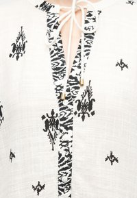 Λευκό μπλουζάκι με υφή, με μαύρα αφηρημένα σχέδια, μπροστινό μέρος με κουμπιά και λαιμόκοψη με δέσιμο που διαθέτει χρυσά μεταλλικά τελειώματα.