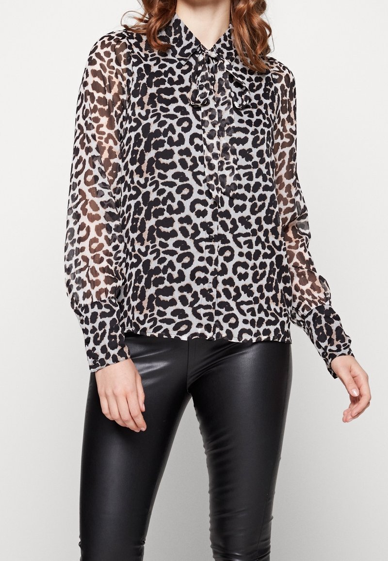 Femme portant un chemisier transparent à imprimé léopard avec manches longues et un nœud au col, assorti à un pantalon en cuir noir.