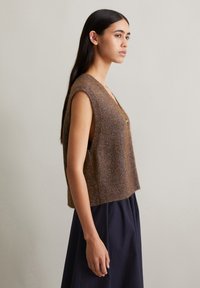 Brown sleeveless knitted vest with a textured finish and a relaxed fit, featuring a V-neck and a ribbed hem. Accented with a central button.