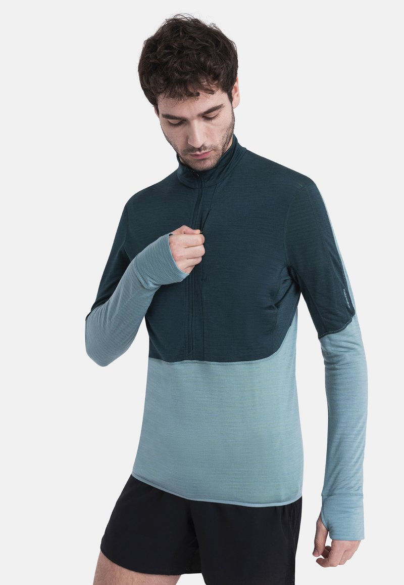 Sudadera de manga larga en verde oscuro y azul claro, hecha de un tejido suave y texturizado, con cremallera de cuarto y orificios para los pulgares en los puños.
