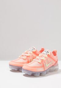 Zapatos Nike Air VaporMax en coral brillante con suela transparente, parte superior de malla y acentos en amarillo neón. Incluye cordones y detalles de marca.