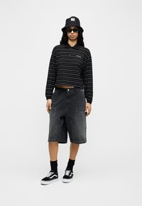 Γυναίκα με μαύρο καπέλο τύπου κάπα, ορθογώνια γυαλιά ηλίου, μαύρο πουλόβερ πόλο με ριγέ μανίκια, ξεβαμμένο μαύρο παντελόνι μέχρι το γόνατο και μαύρα αθλητικά παπούτσια.