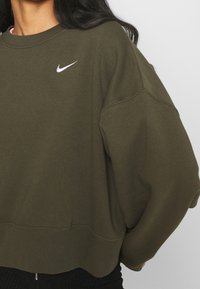 Sudadera verde oliva con un diseño recortado, fabricada en un tejido suave. Presenta un logo de Nike en blanco en el pecho y mangas de corte relajado.