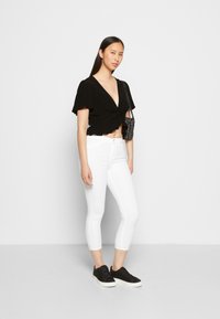 Top preto de manga curta com nó, jeans brancos cropped e ténis pretos. Bolsa texturizada sobre o ombro. Design simples e forma ajustada.