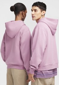 Roza jopa iz mehke tkanine, z raztegljivim krojem in kapucami z zategovalnimi vrvicami. Vidni detajli šivov in večplastna črtasta spodnja majica.