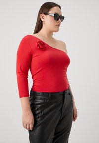 Rød en-skulder topp med roseaccent i myk stoff, kombinert med svarte skinnbukser med frontlommer og slim fit.
