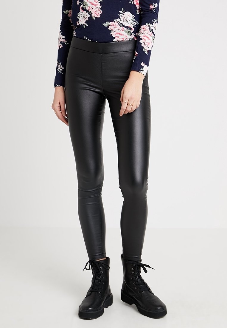 Femme portant un legging noir en simili-cuir, des bottines noires à lacets et un haut à manches longues bleu marine à motif floral, debout devant un fond uni.