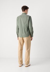 Cămașă verde deschis cu mâneci lungi, purtată cu pantaloni bej. Cămașa are un tiv curbat și o textură netedă.