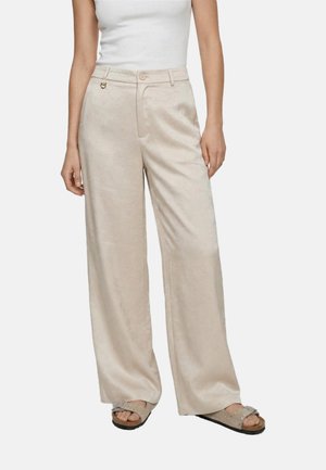 Donna che indossa pantaloni beige a gamba larga, top bianco smanicato e sandali beige slip-on con cinturini e piccole borchie.