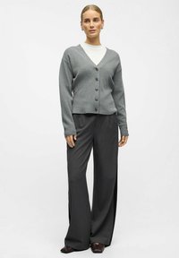 Gray knitted cardigan with a V-neck and six button closure, paired with dark, wide-leg trousers, set against a plain background.