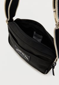 Μαύρη τσάντα χιαστί από καμβά με κλείσιμο με φερμουάρ, διαθέτει λουράκι με λευκές και μαύρες ρίγες και ένα εμφανές σήμα στο μπροστινό μέρος.