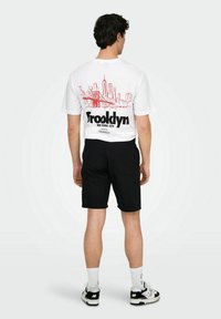 Hvid bomulds t-shirt med s grafik af den røde skyline og teksten "Brooklyn New York City" på ryggen. Sammen med sorte shorts og sneakers.