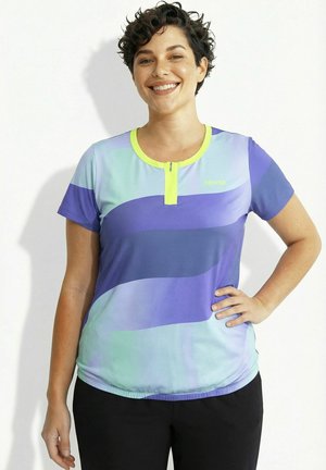 Lächelnde Frau mit kurzen lockigen Haaren, die ein blaues und violettes Sportshirt mit neon-gelbem Rand und schwarze Hosen trägt, steht mit der Hand in der Hüfte.