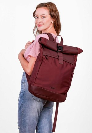 Kaštanově hnědý batoh s rolovacím uzávěrem, černou přezkou, texturovanou tkaninou a pevným dnem. Nastavitelné popruhy a detail s logem vespod.