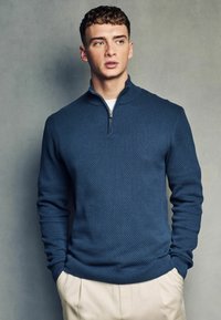 Maglione zip in navy con un design a nido d'ape testurizzato, polsini e orlo a coste, abbinato a pantaloni chiari.