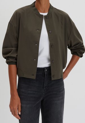 Person wearing a dark olive green buttoned bomber jacket over a white top, paired with black high-waisted jeans, arms relaxed by sides.