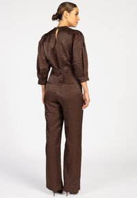 Bruine gestructureerde jumpsuit met een hoge hals, een ceintuur in de taille en een keyhole-detail op de rug. Getailleerde pasvorm met een brede broekspijp en driekwart mouwen.