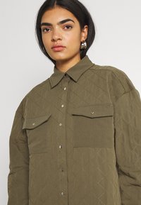 Camisa acolchoada verde-oliva com fecho de botão, dois bolsos no peito e botões de pressão prateados. Padrão em diamante texturizado em toda a peça.