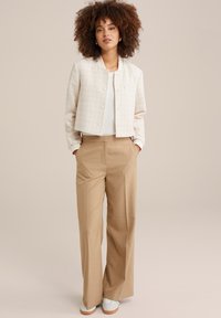 Veste cropped beige clair en tissu texturé, avec fermeture à boutons et un haut blanc. Pantalons larges plissés couleur tan et baskets blanches.
