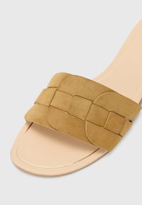 Sandale en suede beige avec une large sangle tissée et une semelle plate. Le pied est léger avec un fin accent doré autour du bord.