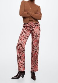 Gilet marron avec boutons associé à un pantalon large à motifs avec des designs paisley rouges et roses sur un fond sombre. Bottes à talons marron.