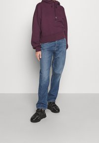 Person som bär en mörk lila kortärmad hoodie, blå jeans och svarta sneakers, stående mot en ljusgrå bakgrund.