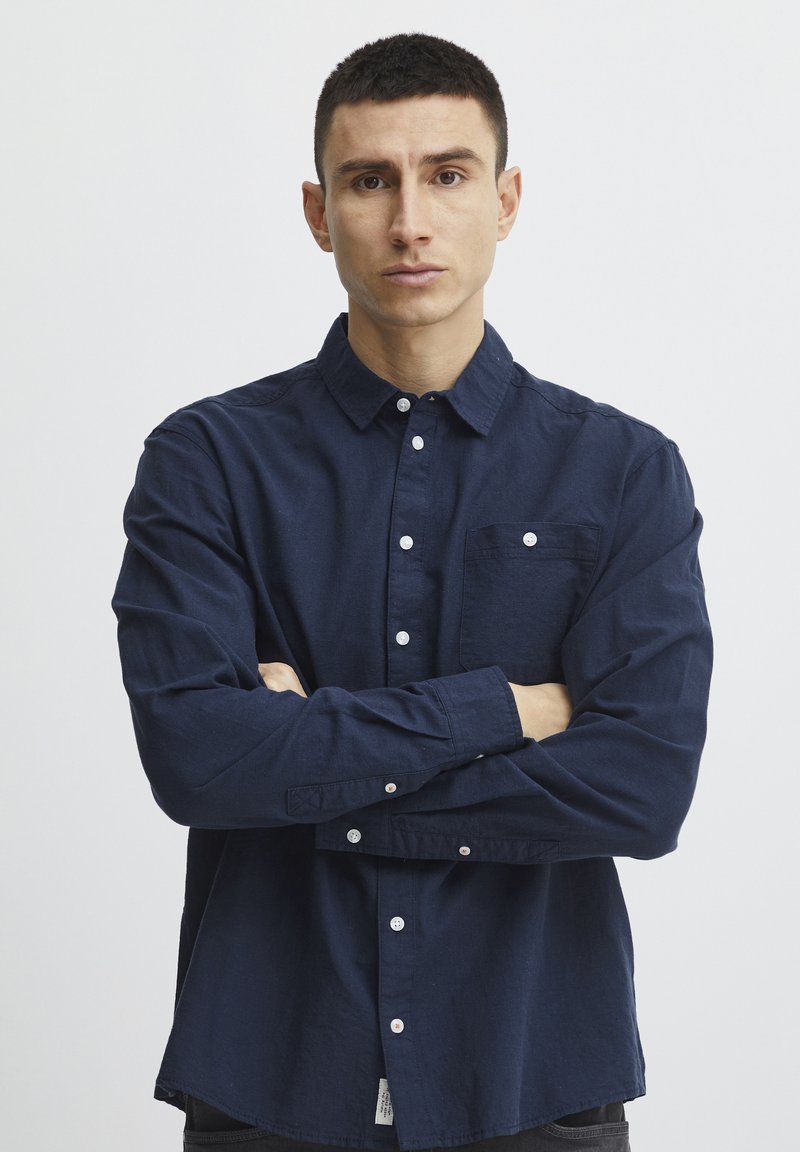 Camicia blu navy con bottoni, maniche lunghe, tasca sul petto e bottoni bianchi. Il tessuto sembra strutturato e leggero.