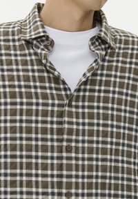 Brązowa i biała koszula w kratkę z kołnierzykiem zapinanym na guziki. Posiada brązowe guziki i gładką teksturę. Noszona na białej koszulce z krótkim rękawem.