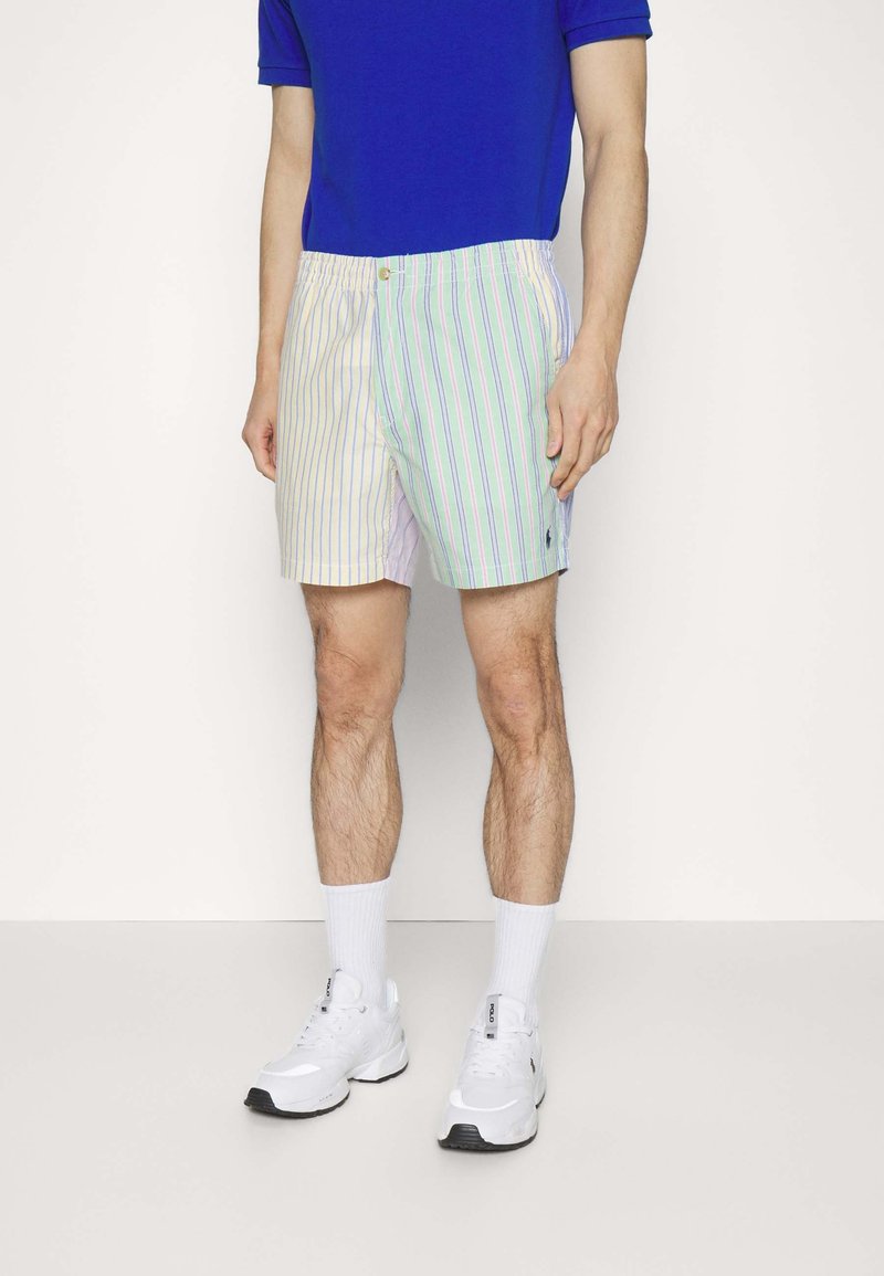 Homme portant un short rayé vertical bicolore jaune pastel et vert avec des baskets blanches et une chemise bleue, debout devant un fond uni.