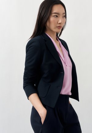 Femme en blazer noir, blouse rose et pantalon noir, debout les mains dans les poches devant un fond clair uni.