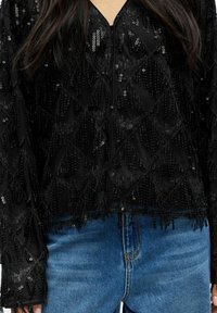 Černá průhledná blůza s černými flitrovými třásněmi, v kombinaci s modrými džíny, odhalující část trupu a část dlouhých tmavých vlasů.