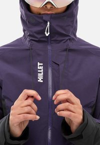 Veste imperméable violette avec col haut, fermeture éclair à l'avant et accents noirs. La marque "MILLET" est imprimée sur la poitrine.
