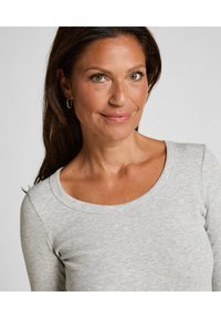 Grijze geribde longsleeve top met een wijde halslijn, met een gladde textuur. Het gezicht van het model is gedeeltelijk zichtbaar en glimlacht zachtjes.