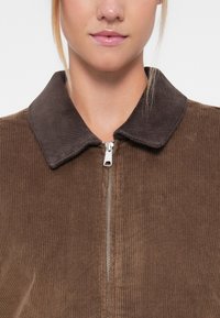 Bruine corduroy jas met een donkerbruine kraag, voorzien van een ritsluiting aan de voorkant en verticale ribbels over de hele jas.