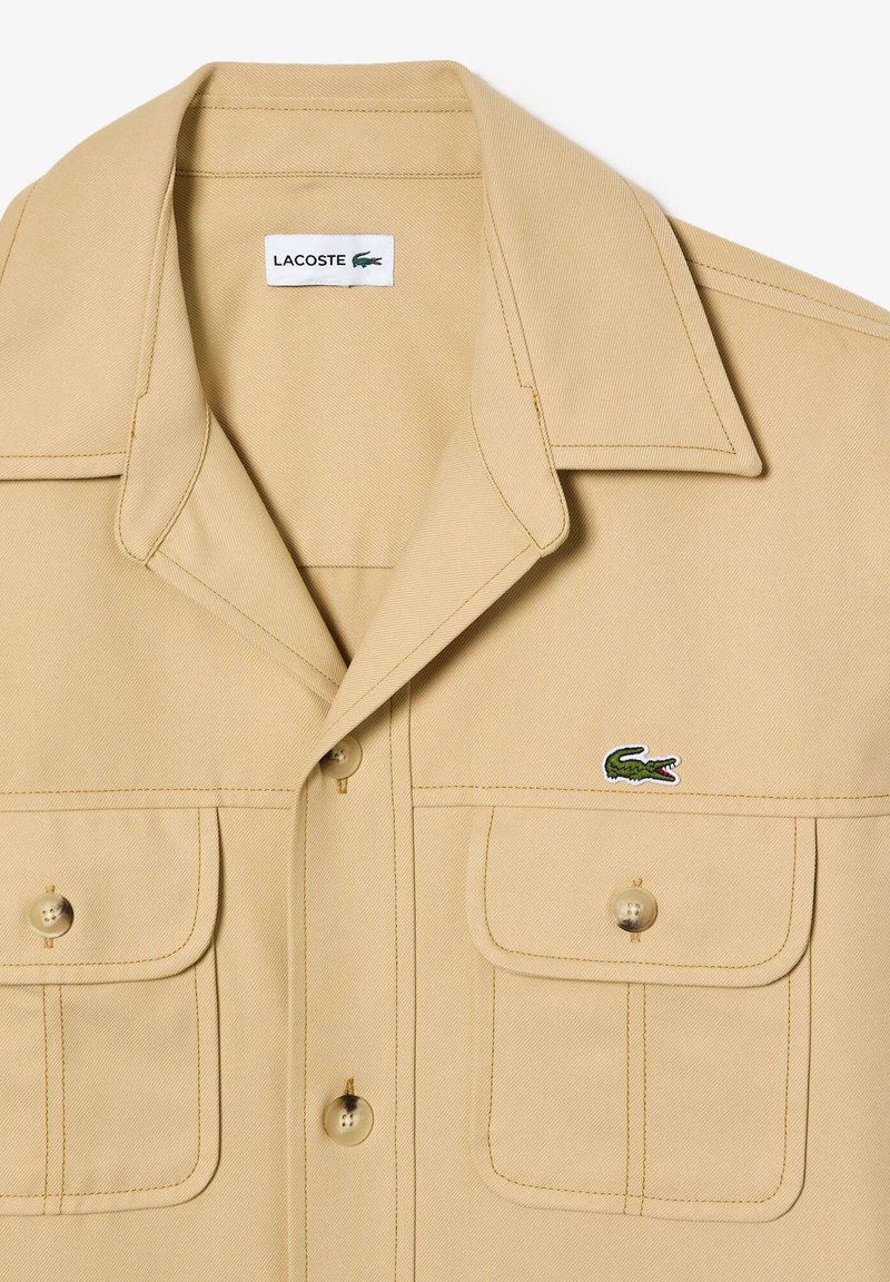 Kaki Knopfleistenhemd mit zwei Brusttaschen, spitzem Kragen und grünem Lacoste-Logo auf der linken Seite. Hergestellt aus strukturiertem Baumwollstoff.