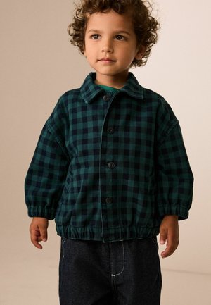 Ungt barn med krøllet hår iført en grøn- og sortternet jakke med knapper og mørke jeans, stående mod en neutral baggrund.