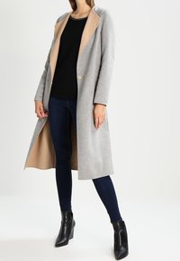 Cappotto grigio in misto lana con revers beige, chiusura a un bottone e maniche lunghe. Indossato sopra un top nero e abbinato a jeans scuri e stivaletti neri.