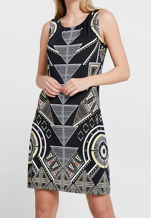 Vrouw in mouwloze zwarte jurk met symmetrische geometrische patronen in wit, geel en roze, staand met gevouwen handen.