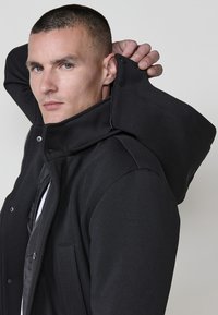Giacca nera con colletto alto e cappuccio oversize, realizzata in tessuto morbido, con zip frontale e tasche laterali.