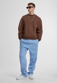 Brun sweatshirt med en afslappet pasform, parret med lys blå sweatpants. Tilbehør inkluderer en sølvkæde og sorte solbriller. Hvide sneakers.