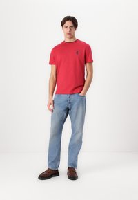 Rød bomull T-skjorte med rund hals og liten mørk logo på brystet, kombinert med lyseblå jeans og brune skinnsko.