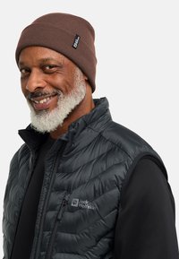 Uomo barbuto sorridente che indossa un berretto lavorato a maglia marrone e un gilet trapuntato nero Jack Wolfskin sopra una maglietta a maniche lunghe nera.
