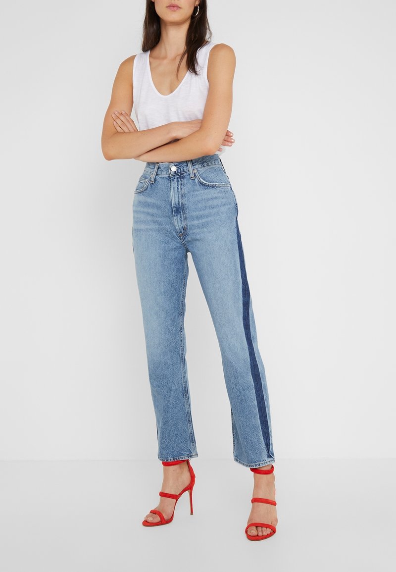 Femme debout, les bras croisés, portant un haut blanc sans manches, un jean bleu taille haute avec une bande latérale foncée et des sandales rouges à talons hauts.