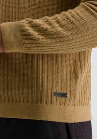 Gestreifter Pullover in Senfgelb mit vertikaler Rippstruktur, ausgestattet mit einem festen Saum und einem markierten Metall-Tag auf der Unterseite.