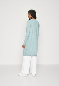 Lys blå lang cardigan med rund halslinje, ribbestrikkede mansjetter og kant. Stylet med hvite vide bukser og hvite joggesko.