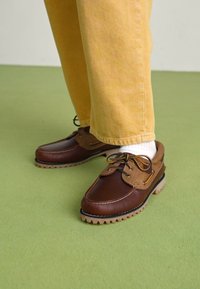 Chaussures en cuir marron avec un accent en cuir beige, à bout rond, à lacets, et semelle en caoutchouc texturée, portées avec un pantalon jaune et des chaussettes blanches.