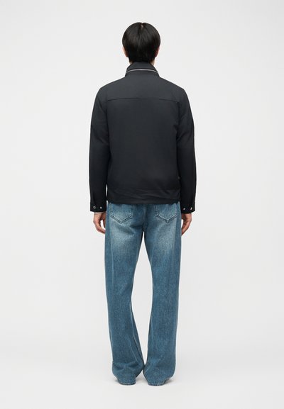 Άνδρας με μαύρο μπουφάν και φαρδύ ανοιχτό γαλάζιο τζιν, που στέκεται γυρισμένος με την πλάτη προς το μέρος μας, πάνω σε λευκό φόντο.