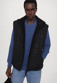 Black sleeveless vest with a textured surface and fleece lining, worn over a blue knitted sweater, paired with light blue jeans.