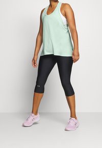 Jasnozielony bezrękawnik sportowy w połączeniu z czarnymi legginsami 3/4 i różowymi butami biegowymi. Teksturowany materiał i krój na plecach w stylu racerback.