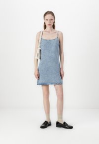 Traper haljina na bretele s tankim trakama, svijetlo plava boja, s rastrganim rubom. Uparena s tamnim cipelama i bež čarapama, uz malu bijelu torbicu.